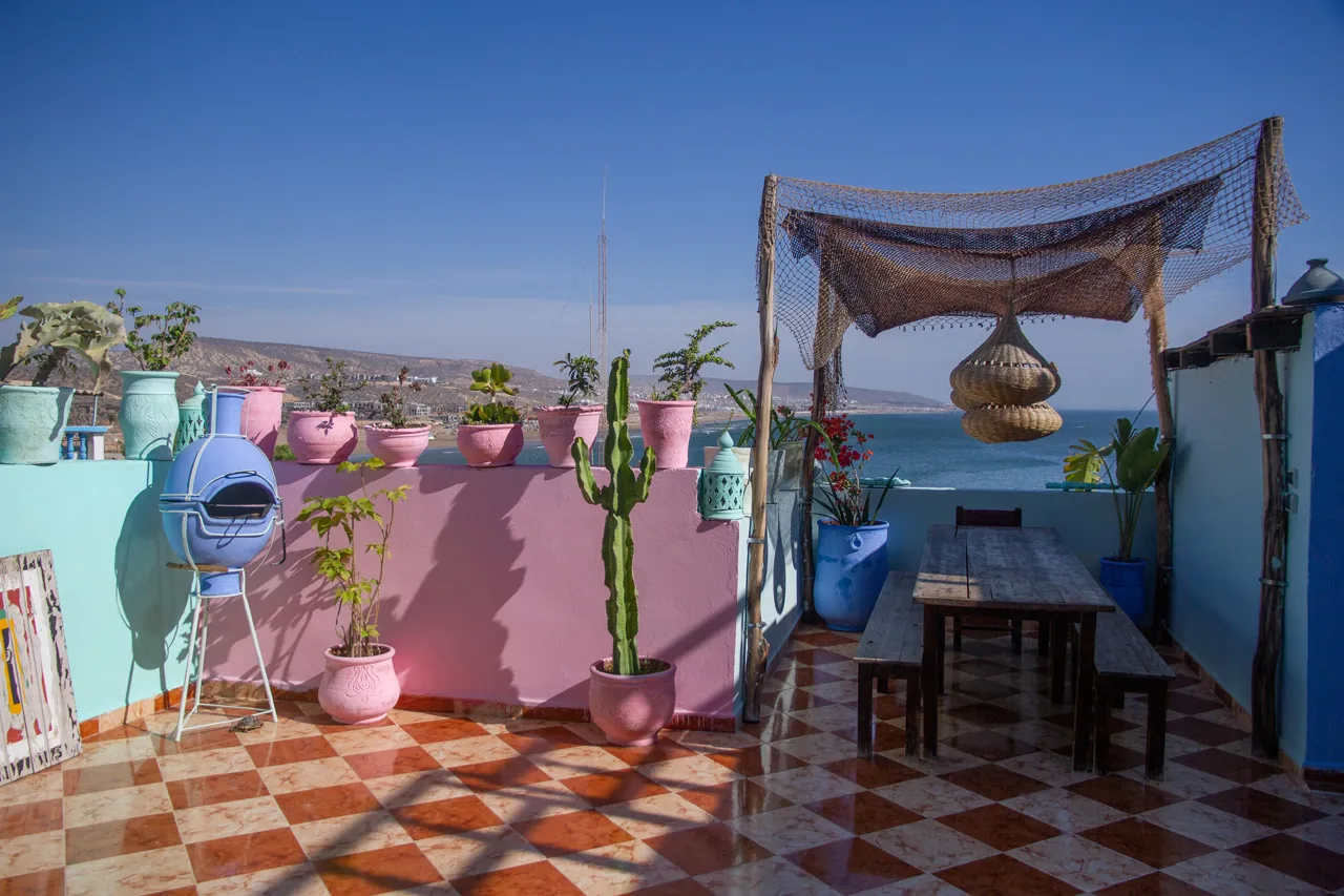 A rooftop café terrace in Tamraght overlooking the Atlantic — surfers in board shorts drinking mint tea after a morning session